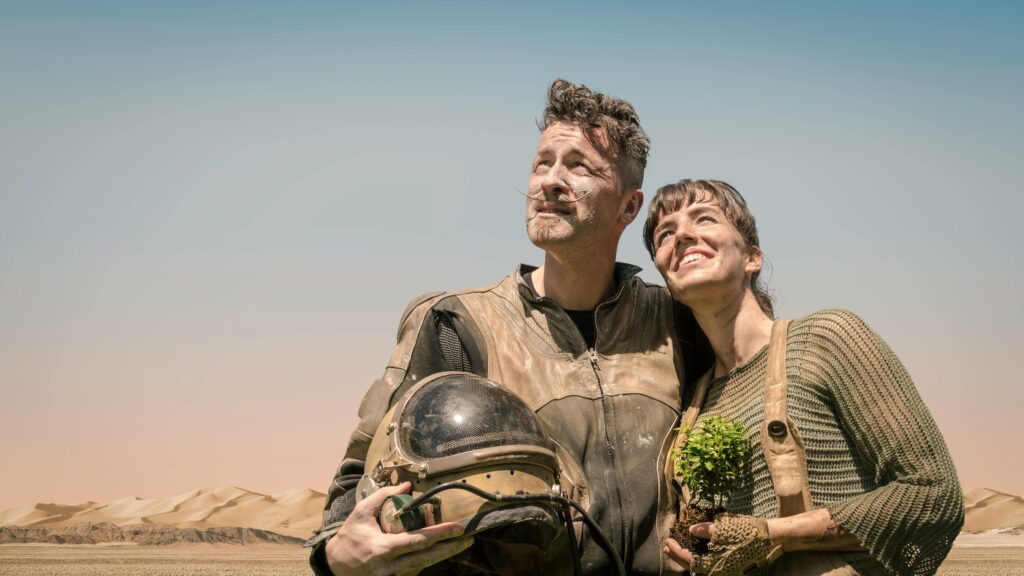 Hombre y mujer en trajes futuristas sosteniendo una planta en un paisaje desértico.