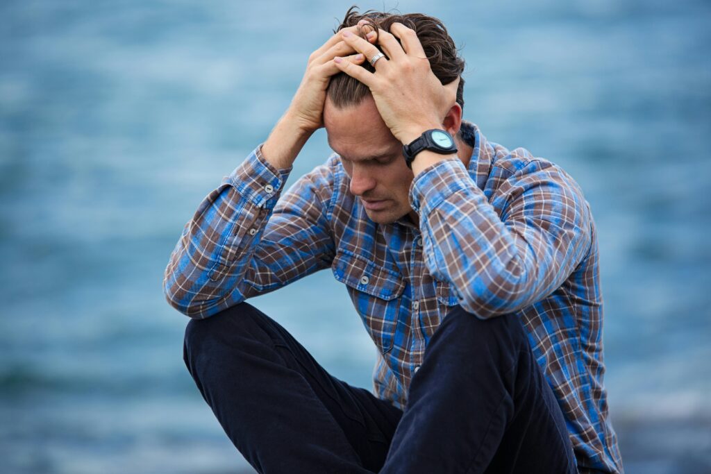 Los peligros ocultos del estrés A man in a plaid shirt sits by the water looking distressed, symbolizing stress.